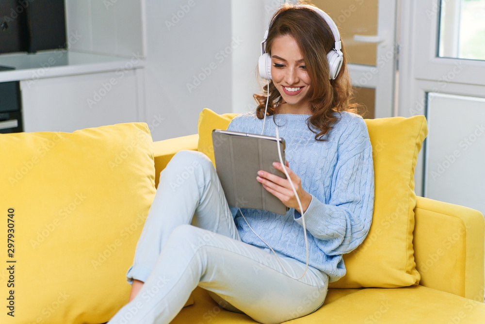 Exuberant 30-aged girl with wavy brown hair wearing white earphones listens music and uses laptop or tablet, resting on the yellow couch at cozy home. Exuberant 30-aged girl with wavy brown hair wearing white earphones listens music and uses laptop or tablet, resting on the yellow couch at cozy home.