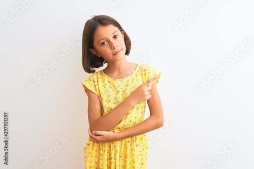 Young beautiful child girl wearing yellow floral dress standing over isolated white background Pointing with hand finger to the side showing advertisement, serious and calm face