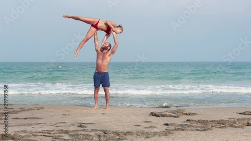 Muscular man and fit young woman training together in Acro Yoga. Morning sun lighting the couple and the mediterranean beach background.