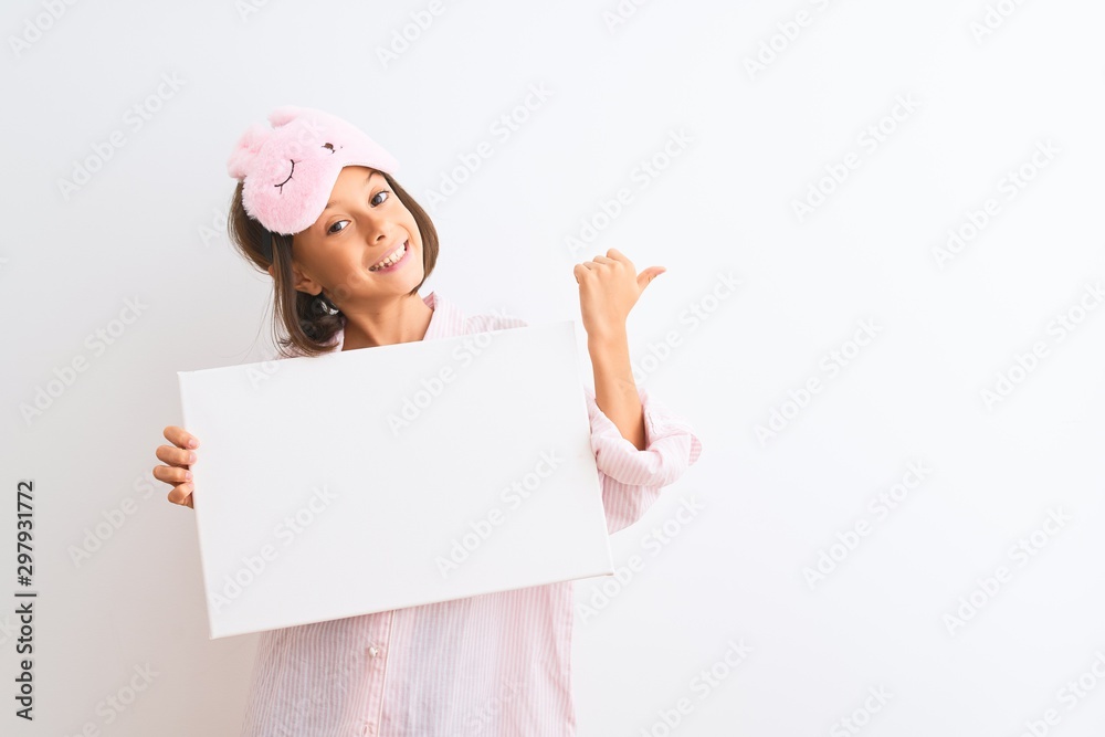 Child girl wearing sleep mask and pajama holding banner over isolated white background pointing and showing with thumb up to the side with happy face smiling Child girl wearing sleep mask and pajama holding banner over isolated white background pointing and showing with thumb up to the side with happy face smiling