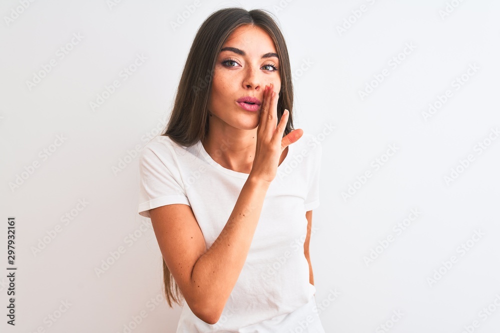 Young beautiful woman wearing casual t-shirt standing over isolated white background hand on mouth telling secret rumor, whispering malicious talk conversation Young beautiful woman wearing casual t-shirt standing over isolated white background hand on mouth telling secret rumor, whispering malicious talk conversation