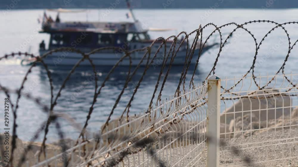 A fence with barbed wire on background a sailing ship in the Mediterranean Sea. Guarded restricted area. Border on the coast A fence with barbed wire on background a sailing ship in the Mediterranean Sea. Guarded restricted area. Border on the coast