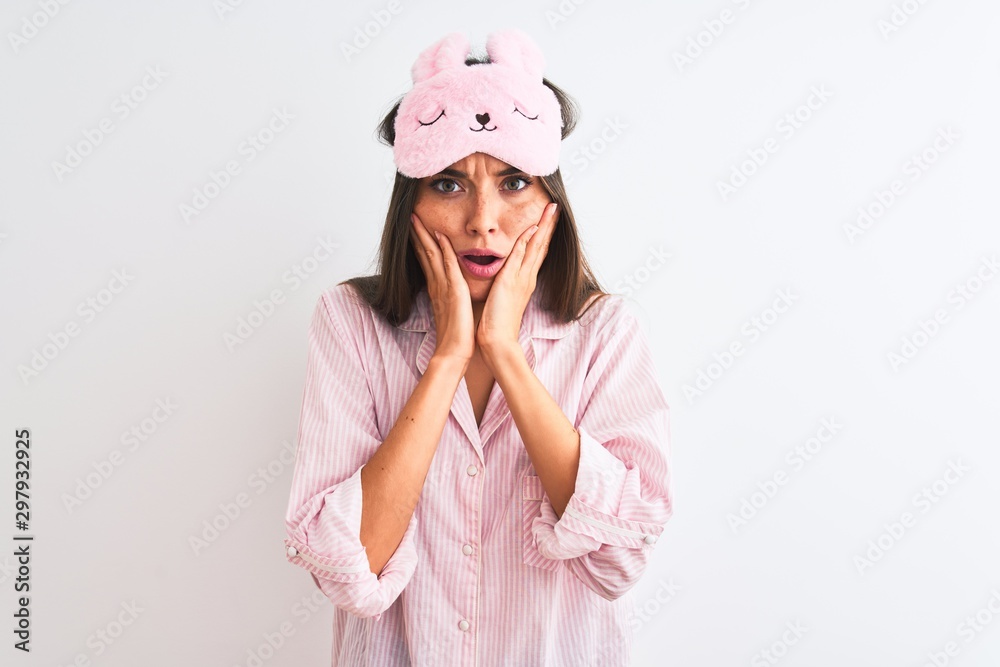 Young beautiful woman wearing sleep mask and pajama over isolated white background afraid and shocked, surprise and amazed expression with hands on face Young beautiful woman wearing sleep mask and pajama over isolated white background afraid and shocked, surprise and amazed expression with hands on face