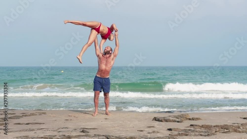 Muscular man and fit young woman training together in Acro Yoga. Morning sun lighting the couple and the mediterranean beach background.