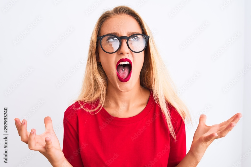 Young beautiful woman wearing red t-shirt and glasses standing over isolated white background crazy and mad shouting and yelling with aggressive expression and arms raised. Frustration concept. Young beautiful woman wearing red t-shirt and glasses standing over isolated white background crazy and mad shouting and yelling with aggressive expression and arms raised. Frustration concept.