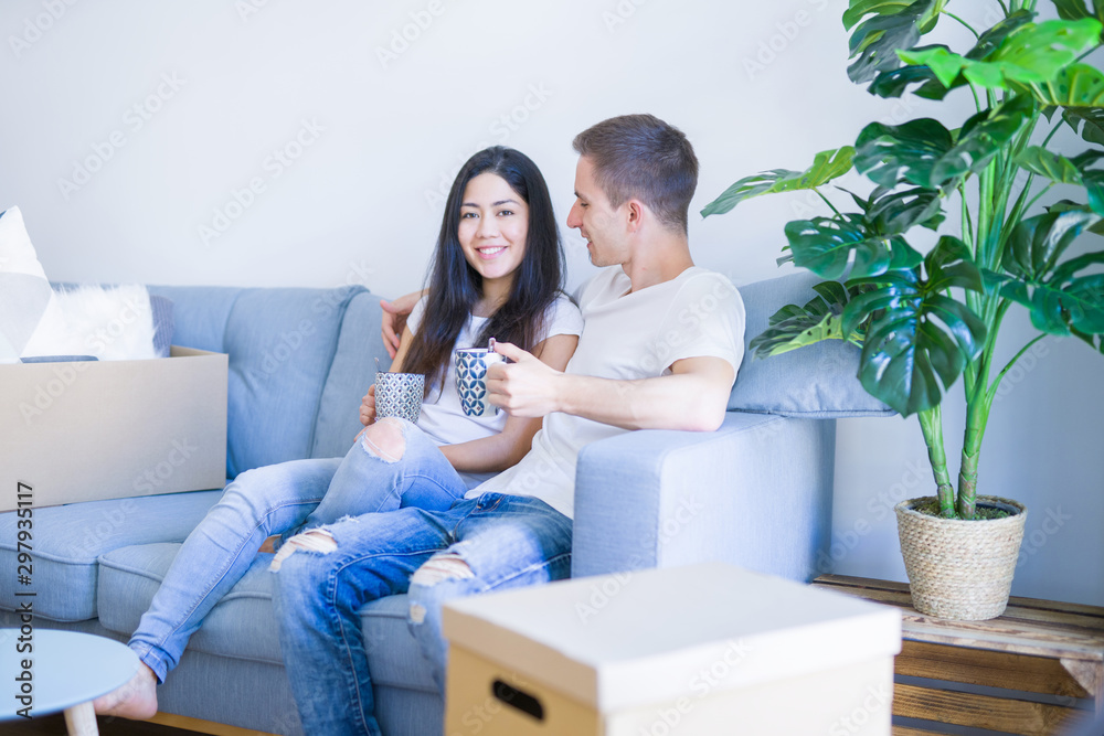 Young beautiful couple sitting on the sofa drinking coffee at new home around cardboard boxes Young beautiful couple sitting on the sofa drinking coffee at new home around cardboard boxes
