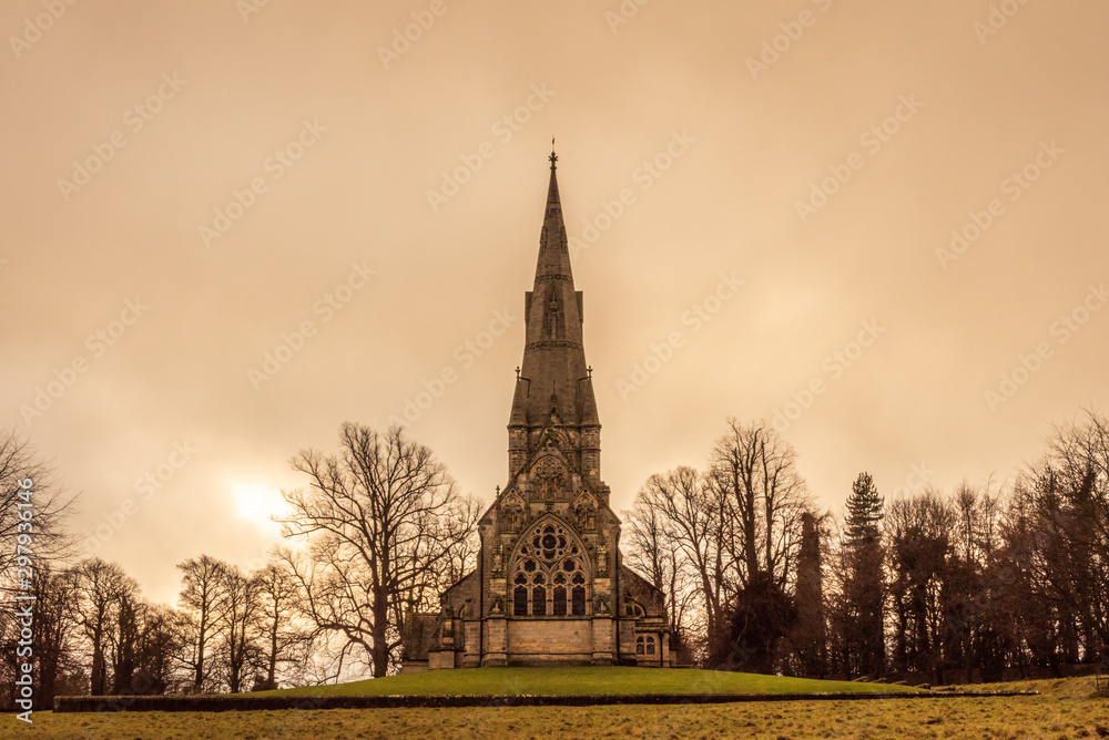 Obraz premium A beautiful and gothic church situated in a park with winter trees all around during a dramatic and moody afternoon in Studley Park, UK.