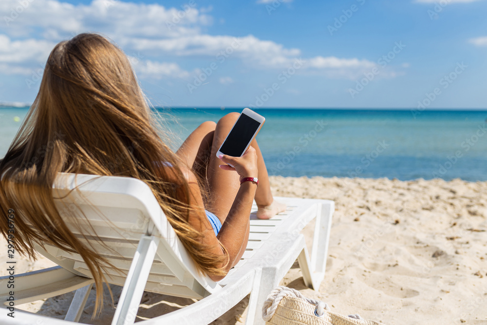 Young girl lying on beach with mobile phone in hand on tropical island. Girl on vacation at sea uses a smartphone and social networks on the background of sea waves
