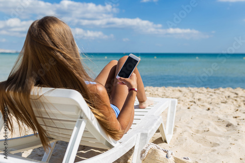 Young girl lying on beach with mobile phone in hand on tropical island. Girl on vacation at sea uses a smartphone and social networks on the background of sea waves