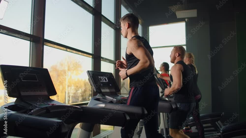 Two men running on a treadmill in the background of a large window in the fitness room. Young men running in the fitness room. Aerobic workouts in the gym
