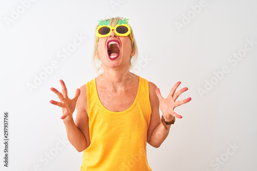 Middle age woman on vacation wearing pineapple sunglasses over isolated white background crazy and mad shouting and yelling with aggressive expression and arms raised. Frustration concept.