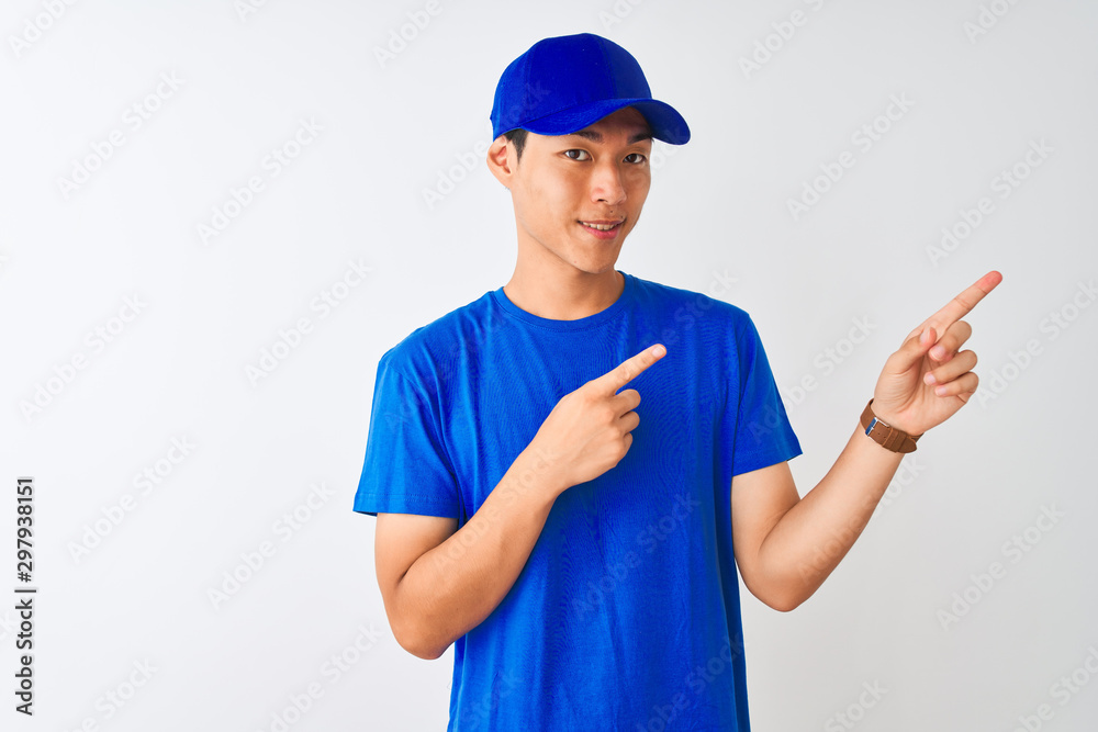 Chinese deliveryman wearing blue t-shirt and cap standing over isolated white background smiling and looking at the camera pointing with two hands and fingers to the side.