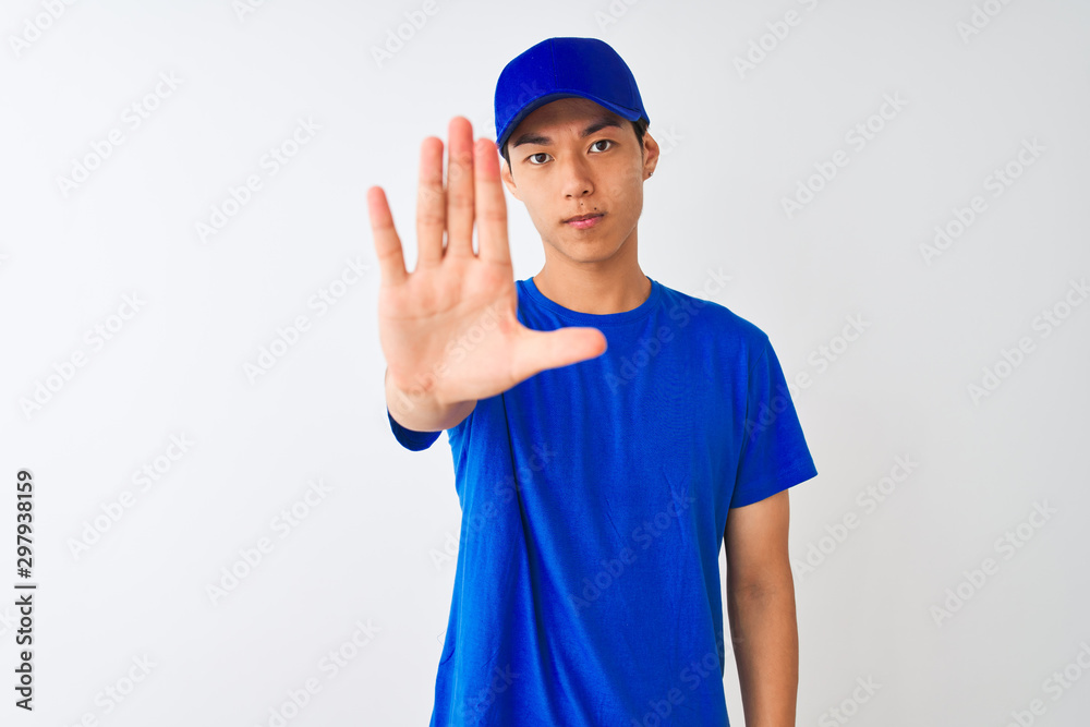 Chinese deliveryman wearing blue t-shirt and cap standing over isolated white background doing stop sing with palm of the hand. Warning expression with negative and serious gesture on the face.
