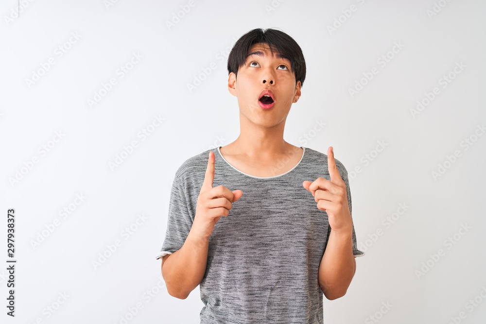 Young chinese man wearing casual t-shirt standing over isolated white background amazed and surprised looking up and pointing with fingers and raised arms. Young chinese man wearing casual t-shirt standing over isolated white background amazed and surprised looking up and pointing with fingers and raised arms.
