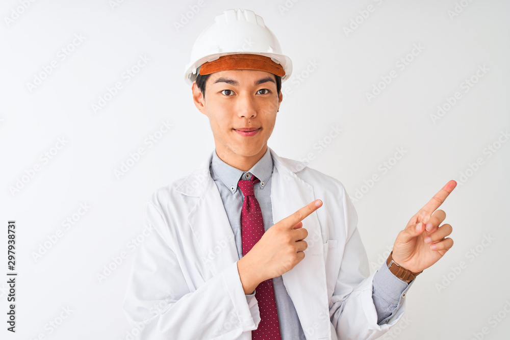 Chinese architect man wearing coat and helmet standing over isolated white background smiling and looking at the camera pointing with two hands and fingers to the side.