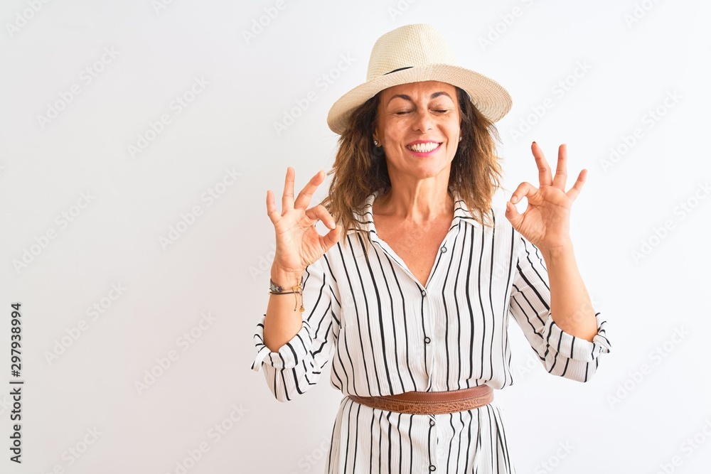 Middle age businesswoman wearing striped dress and hat over isolated white background relax and smiling with eyes closed doing meditation gesture with fingers. Yoga concept. Middle age businesswoman wearing striped dress and hat over isolated white background relax and smiling with eyes closed doing meditation gesture with fingers. Yoga concept.