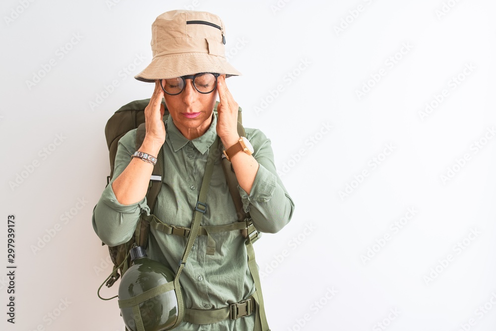 Middle age hiker woman wearing backpack canteen hat glasses over isolated white background with hand on head for pain in head because stress. Suffering migraine. Middle age hiker woman wearing backpack canteen hat glasses over isolated white background with hand on head for pain in head because stress. Suffering migraine.
