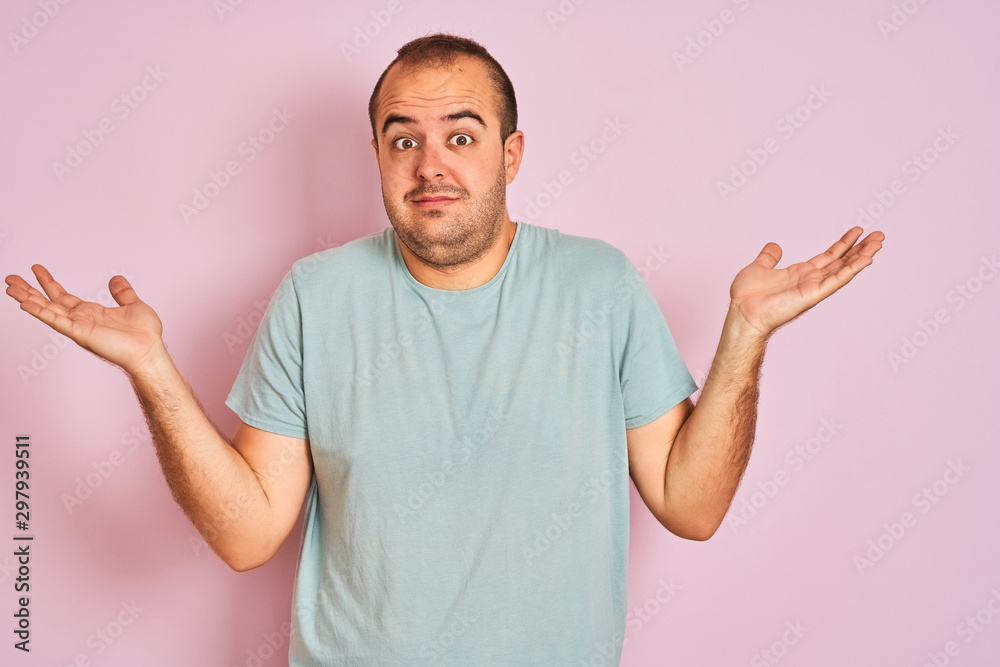 Young man wearing blue casual t-shirt standing over isolated pink background clueless and confused expression with arms and hands raised. Doubt concept. Young man wearing blue casual t-shirt standing over isolated pink background clueless and confused expression with arms and hands raised. Doubt concept.
