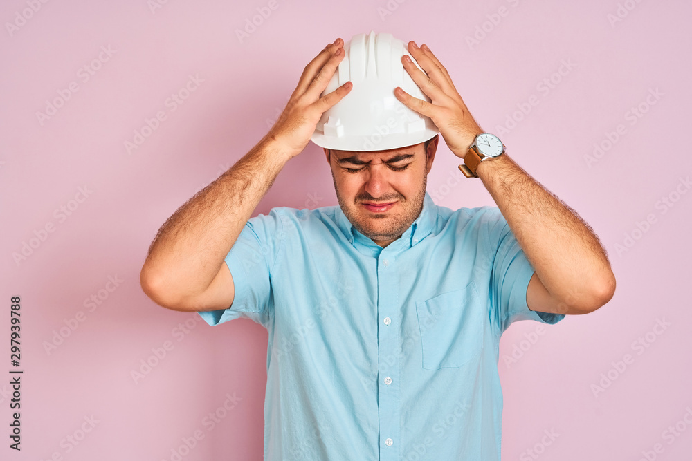 Young architect man wearing security helmet standing over isolated pink background suffering from headache desperate and stressed because pain and migraine. Hands on head.
