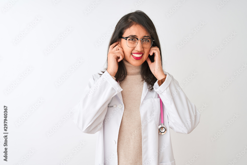 Chinese doctor woman wearing coat and pink stethoscope over isolated white background covering ears with fingers with annoyed expression for the noise of loud music. Deaf concept. Chinese doctor woman wearing coat and pink stethoscope over isolated white background covering ears with fingers with annoyed expression for the noise of loud music. Deaf concept.