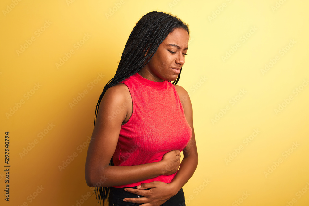 African american woman wearing red casual t-shirt standing over isolated yellow background with hand on stomach because nausea, painful disease feeling unwell. Ache concept. African american woman wearing red casual t-shirt standing over isolated yellow background with hand on stomach because nausea, painful disease feeling unwell. Ache concept.