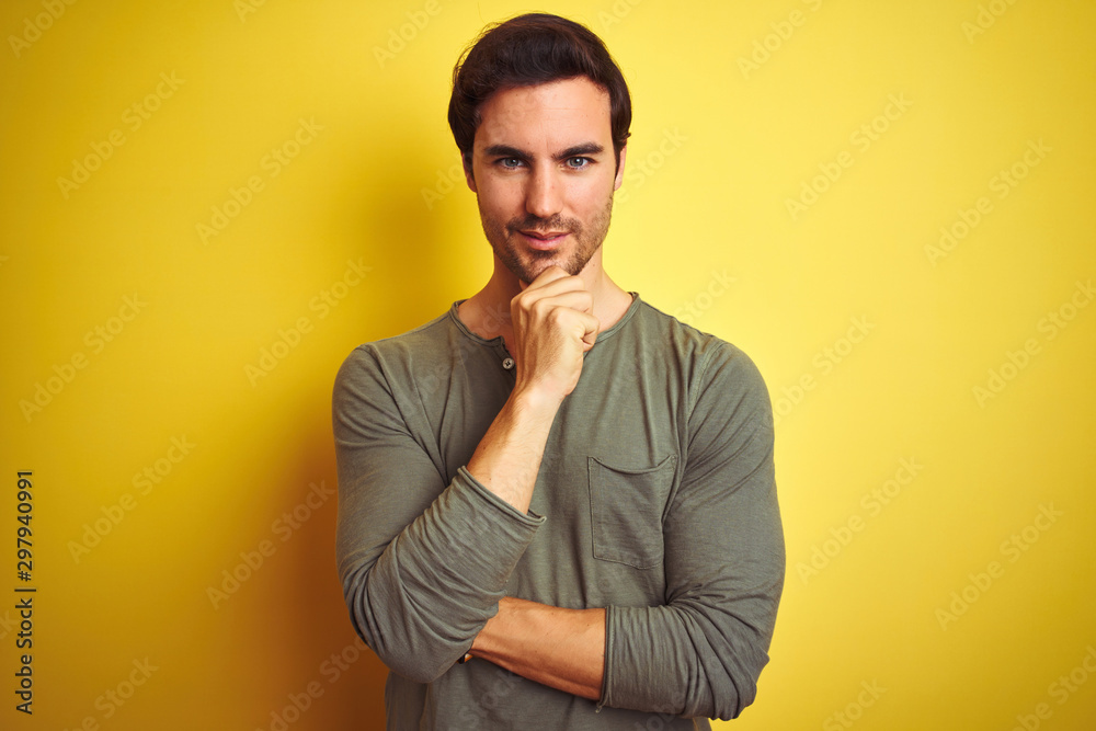 Young handsome man wearing casual t-shirt standing over isolated yellow background looking confident at the camera with smile with crossed arms and hand raised on chin. Thinking positive.