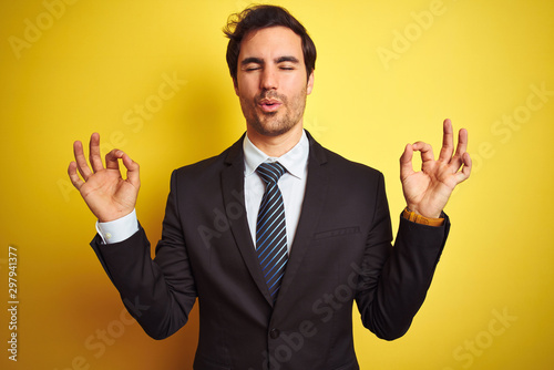 Young handsome businessman wearing suit and tie standing over isolated yellow background relaxed and smiling with eyes closed doing meditation gesture with fingers. Yoga concept.