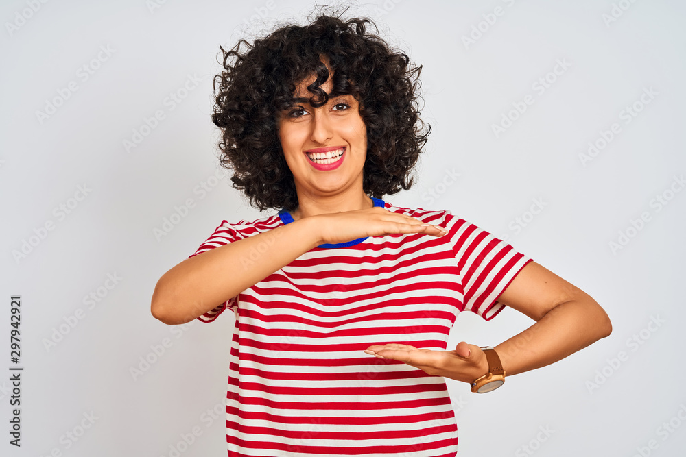 Young arab woman with curly hair wearing striped t-shirt over isolated white background gesturing with hands showing big and large size sign, measure symbol. Smiling looking at the camera. Young arab woman with curly hair wearing striped t-shirt over isolated white background gesturing with hands showing big and large size sign, measure symbol. Smiling looking at the camera.
