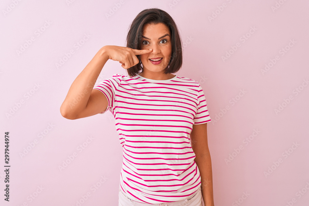 Young beautiful woman wearing striped t-shirt standing over isolated pink background Pointing with hand finger to face and nose, smiling cheerful. Beauty concept Young beautiful woman wearing striped t-shirt standing over isolated pink background Pointing with hand finger to face and nose, smiling cheerful. Beauty concept