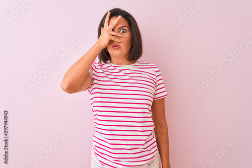Young beautiful woman wearing striped t-shirt standing over isolated pink background peeking in shock covering face and eyes with hand  looking through fingers with embarrassed expression.