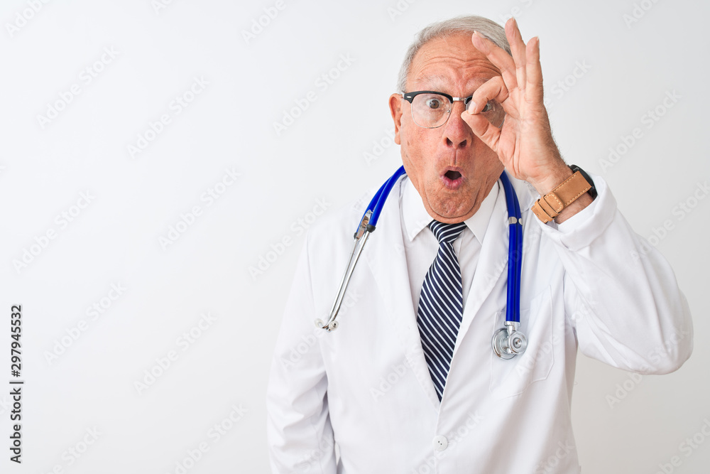 Senior grey-haired doctor man wearing stethoscope standing over isolated white background doing ok gesture shocked with surprised face, eye looking through fingers. Unbelieving expression. Senior grey-haired doctor man wearing stethoscope standing over isolated white background doing ok gesture shocked with surprised face, eye looking through fingers. Unbelieving expression.