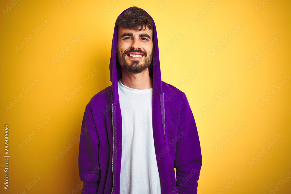 Young man with tattoo wearing purple sweatshirt with hood over isolated yellow background with a happy and cool smile on face. Lucky person. Young man with tattoo wearing purple sweatshirt with hood over isolated yellow background with a happy and cool smile on face. Lucky person.