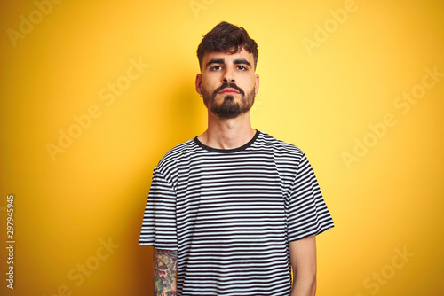 Young man with tattoo wearing striped t-shirt standing over isolated yellow background with serious expression on face. Simple and natural looking at the camera.