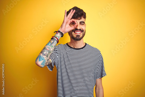 Young man with tattoo wearing striped t-shirt standing over isolated yellow background doing ok gesture with hand smiling, eye looking through fingers with happy face.