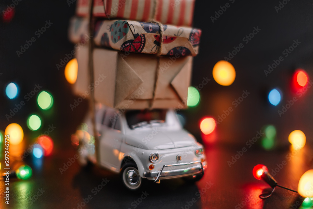 Toy retro car with gift boxes on the roof tied with jute rope. Blurred multicolored lights of garland in the dark background. Happy New Year mood. Merry Christmas and holidays concept. Selective focus