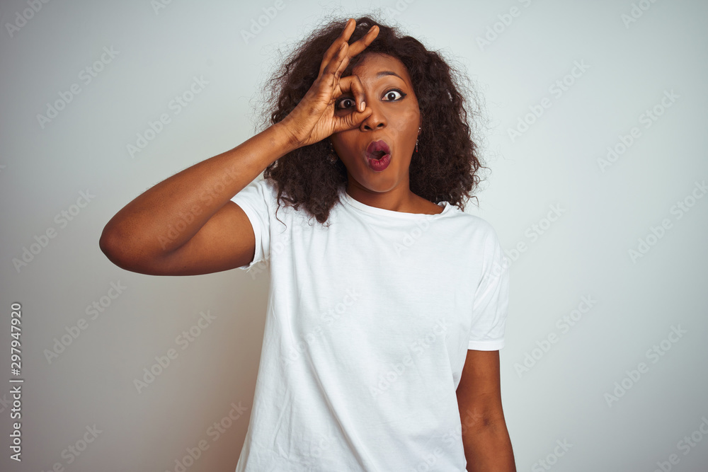 Young african american woman wearing t-shirt standing over isolated white background doing ok gesture shocked with surprised face, eye looking through fingers. Unbelieving expression. Young african american woman wearing t-shirt standing over isolated white background doing ok gesture shocked with surprised face, eye looking through fingers. Unbelieving expression.
