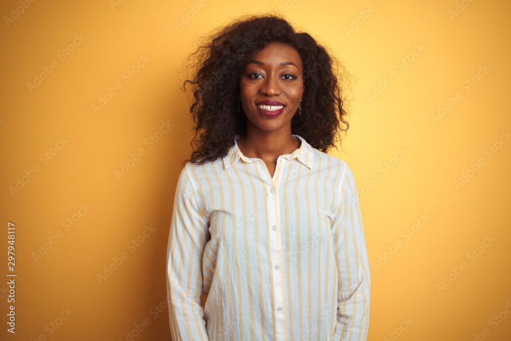 African american woman wearing striped shirt standing over isolated yellow background with a happy and cool smile on face. Lucky person. African american woman wearing striped shirt standing over isolated yellow background with a happy and cool smile on face. Lucky person.