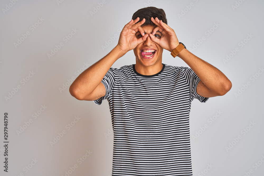 Young indian man wearing navy striped t-shirt standing over isolated white background doing ok gesture like binoculars sticking tongue out, eyes looking through fingers. Crazy expression. Young indian man wearing navy striped t-shirt standing over isolated white background doing ok gesture like binoculars sticking tongue out, eyes looking through fingers. Crazy expression.