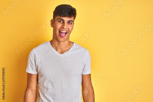 Young indian man wearing white t-shirt standing over isolated yellow background sticking tongue out happy with funny expression. Emotion concept.