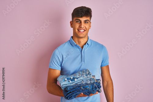 Young indian shopkeeper man holding folded jeans standing over isolated pink background with a happy face standing and smiling with a confident smile showing teeth