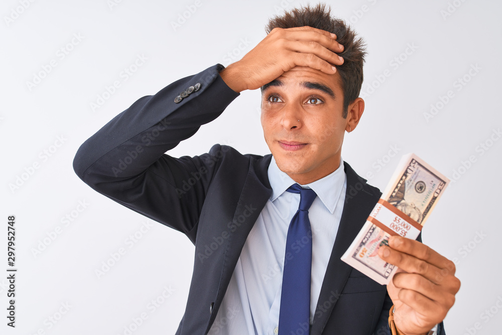 Young handsome businessman wearing suit holding dollars over isolated white background stressed with hand on head, shocked with shame and surprise face, angry and frustrated. Fear and upset Young handsome businessman wearing suit holding dollars over isolated white background stressed with hand on head, shocked with shame and surprise face, angry and frustrated. Fear and upset
