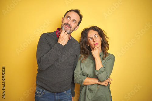 Beautiful middle age couple over isolated yellow background with hand on chin thinking about question, pensive expression. Smiling with thoughtful face. Doubt concept.