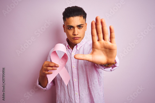 Young brazilian man holding cancer ribbon standing over isolated pink background with open hand doing stop sign with serious and confident expression, defense gesture © Krakenimages.com
