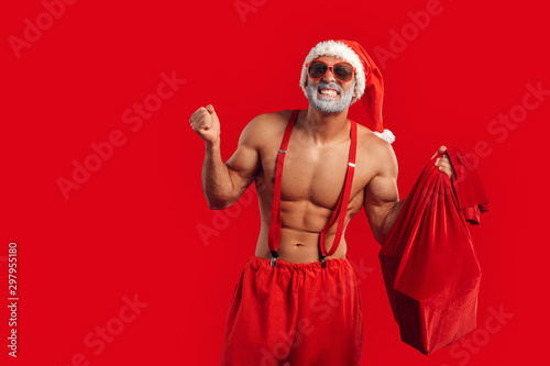 Christmas Freestyle. Young bearded Santa Claus bare muscular upper body in hat and sunglasses standing isolated on red with gift bag hands aside smiling toothy