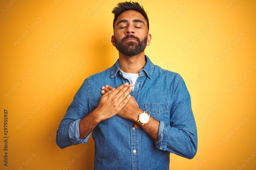 Young indian man wearing denim shirt standing over isolated yellow background smiling with hands on chest with closed eyes and grateful gesture on face. Health concept. Young indian man wearing denim shirt standing over isolated yellow background smiling with hands on chest with closed eyes and grateful gesture on face. Health concept.