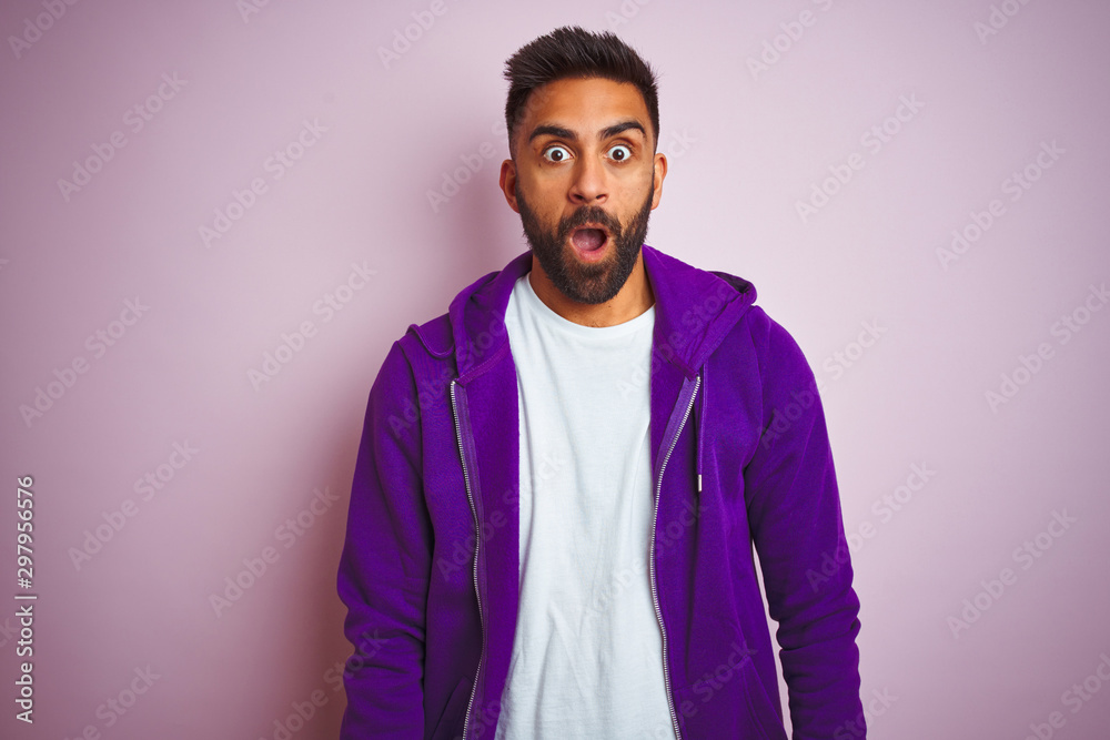 Young indian man wearing purple sweatshirt standing over isolated pink background afraid and shocked with surprise expression, fear and excited face. Young indian man wearing purple sweatshirt standing over isolated pink background afraid and shocked with surprise expression, fear and excited face.