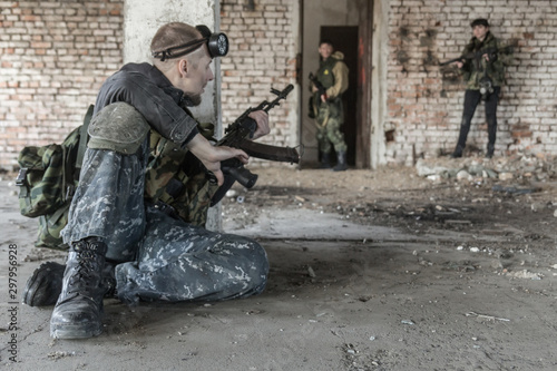 A military man in camo clothes (soldier of fortune) during an exchange of fire hides from enemies behind a column in an abandoned building. In his hands he holds a Kalashnikov assault rifle (AK-74).