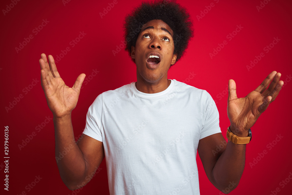 Young american man with afro hair wearing white t-shirt standing over isolated red background crazy and mad shouting and yelling with aggressive expression and arms raised. Frustration concept. Young american man with afro hair wearing white t-shirt standing over isolated red background crazy and mad shouting and yelling with aggressive expression and arms raised. Frustration concept.