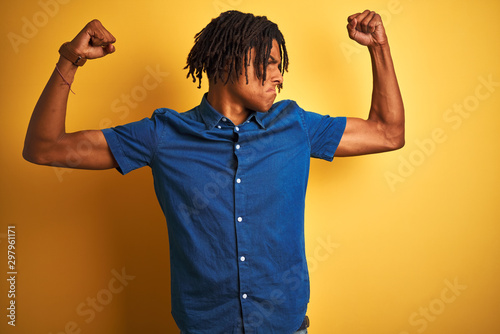 Afro man with dreadlocks wearing casual denim shirt standing over isolated yellow background showing arms muscles smiling proud. Fitness concept.