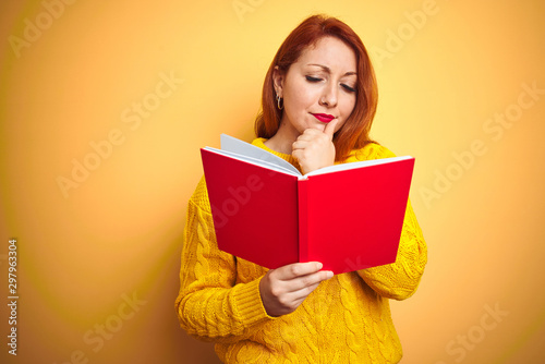 Young redhead teacher woman reading red book over yellow isolated background serious face thinking about question, very confused idea